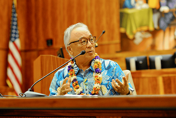 Eric Yamamoto, wearing a blue and white floral shirt and floral lei, discusses the importance of having a federal court vacate Korematsu’s conviction, some 40 years later.