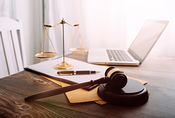 Gold scales of justice sitting on a table with a laptop, pen, paper and judge's gavel.